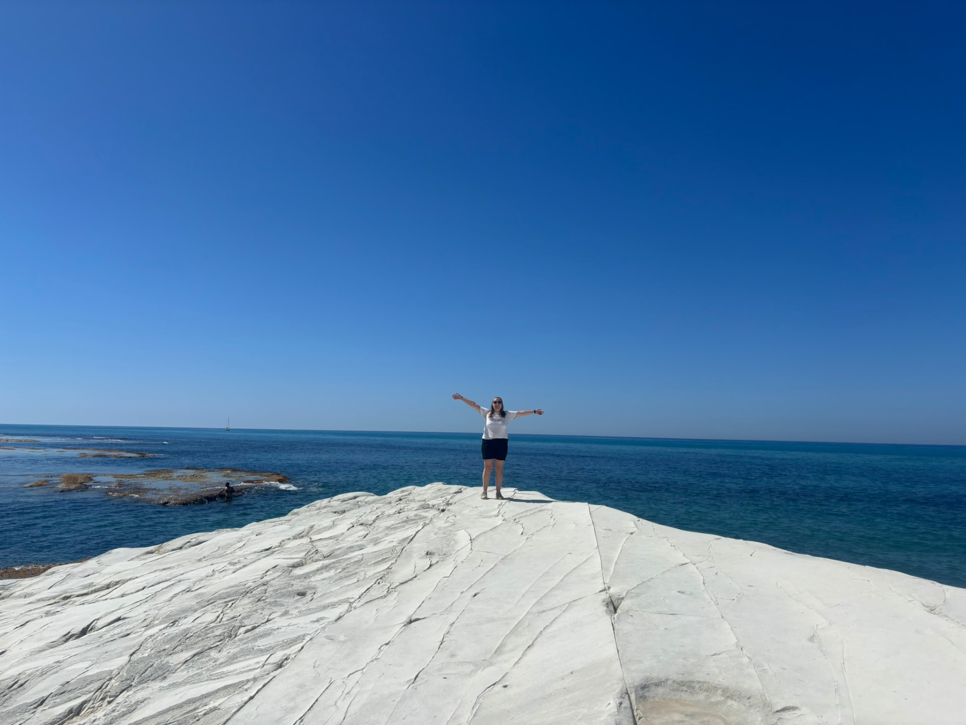 A person standing on a white rock formation overlooking the sea under a clear blue sky